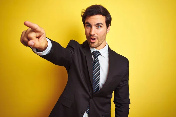 スーツとネクタイを着た若いハンサムなビジネスマンが孤立した黄色の背景の上に立って指を指し 口を開いて驚いた表情 前面に何か — ストック写真