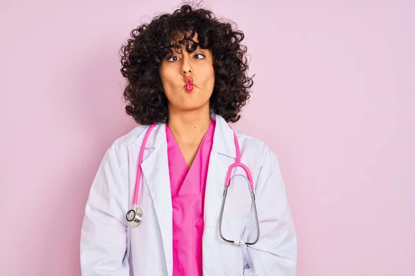 Young arab doctor woman with curly hair wearing stethoscope over isolated pink background making fish face with lips, crazy and comical gesture. Funny expression.