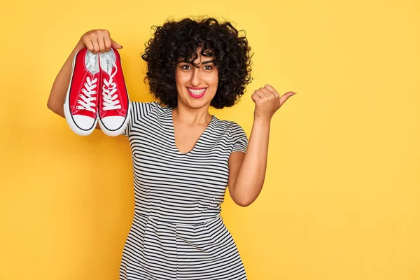Kıvırcık saçlı genç arap kadın izole sarı arka plan üzerinde rahat spor ayakkabı tutan işaret ve mutlu yüzü gülümseyerek yan başparmak ile gösteren