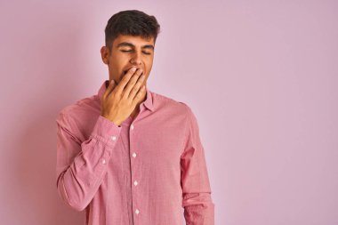 İzole pembe arka plan üzerinde duran zarif gömlek giyen genç hintli adam el ile ağız kapsayan yorgun esneme sıkılmış. Huzursuz ve uykululuk.