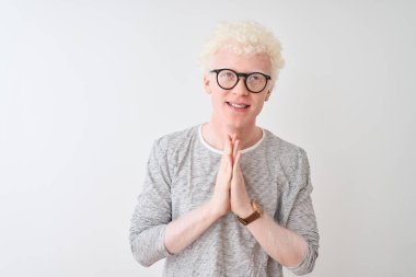 Genç albino sarışın adam çizgili t-shirt ve gözlük izole beyaz arka plan üzerinde elleri birlikte emin gülümseyerek af isteyen dua giyiyor.