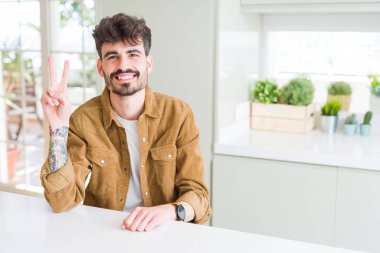 Beyaz masanın üzerinde oturan rahat ceket giyen genç adam, kendinden emin ve mutlu gülümserken iki numaralı parmaklarıyla işaret ediyor..