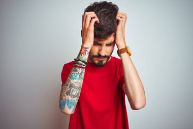 Kırmızı t-shirt giyen dövme giyen genç adam baş ağrısı umutsuz muzdarip izole beyaz arka plan üzerinde duran ve stresli çünkü ağrı ve migren. Eller baş üstünde.