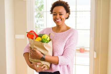 Kağıt torba taze yiyecek tam ile ayakta ve diş gösterilen kendine güvenen bir gülümsemeyle gülümseyerek mutlu bir yüz tutan genç Afro-Amerikan kadın