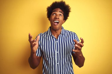 Afro saçlı Amerikalı adam çizgili gömlek giyen izole sarı arka plan üzerinde duran deli ve deli bağırarak ve agresif ifade ve kollar kaldırdı ile bağırıyor. Hayal kırıklığı kavramı.