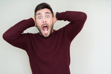 Genç yakışıklı adam izole arka plan üzerinde bir kazak giyen Crazy ve baş elleri ile korkmuş, korkmuş ve açık ağız ile şok şaşırttı