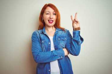 Beyaz izole arka plan üzerinde duran denim gömlek giyen genç güzel kızıl saçlı kadın zafer işareti yapıyor kameraya göz kırparak mutlu yüz ile gülümseyerek. İki numara.