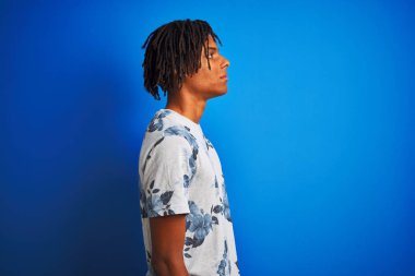 Yan bakarak izole mavi arka plan üzerinde yaz çiçek t-shirt giyen dreadlocks ile Afro amerikan adam, kendine güvenen gülümseme ile doğal yüzü ile profil poz dinlenmek.