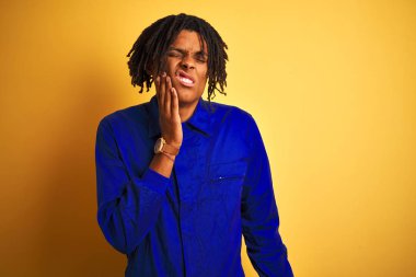 Dreadlocks giyen dreadlocks ile Afro işçi adam izole sarı arka plan üzerinde diş ağrısı veya diş üzerinde diş hastalığı nedeniyle ağrılı ifade ile el ile ağız dokunmadan mekanik üniforma giyiyor. Diş hekimi konsepti.