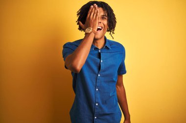 El ile bir göz kapsayan izole sarı arka plan üzerinde duran rahat kot gömlek giyen dreadlocks giyen Afro adam, yüzünde kendinden emin gülümseme ve sürpriz duygu.