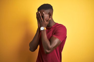 Kırmızı tişört giyen genç afro-amerikan adam izole sarı arka plan üzerinde ayakta üzgün ifade ile elleri ile yüz kapsayan ağlarken. Depresyon kavramı.