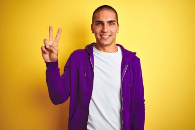Genç yakışıklı adam sarı izole arka plan üzerinde mor sweatshirt giyen mutlu yüz zafer işareti yapıyor kameraya göz kırparak gülümseyerek. İki numara.