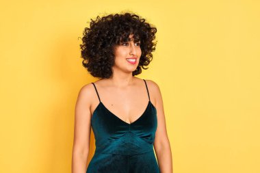Kıvırcık saçlı genç arap kadın izole sarı arka plan üzerinde zarif elbise giyen yüzünde gülümseme, doğal ifade ile yan bakıyor. Gülmekten emin.