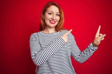 Genç kızıl saçlı kadın kırmızı izole arka plan üzerinde duran kayışlar lacivert gömlek giyen gülümseyerek ve yan iki el ve parmakları ile işaret kamera bakıyor.