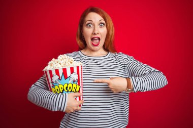 Genç güzel kızıl saçlı kadın kırmızı izole arka plan üzerinde patlamış mısır yeme çok mutlu el ve parmak ile işaret