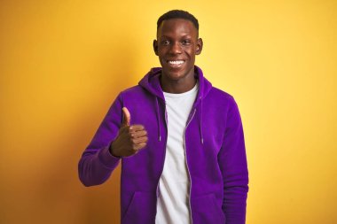 Izole sarı arka plan üzerinde duran mor sweatshirt giyen Afro-Amerikan adam el ile jest kadar mutlu başparmak yapıyor. Başarı göstererek kameraya bakan ifadeyi onaylama.