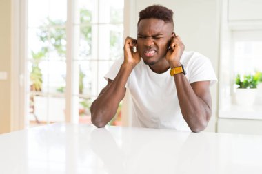 Yakışıklı afro-amerikan adam evde beyaz masada yüksek sesle müzik sesi için rahatsız ifade ile parmakları ile kulakları kapsayan. Sağır kavramı.