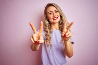 Mor tişört giyen genç güzel kadın pembe izole arka plan üzerinde duran gülümseyerek kameraya bakarak parmakları zafer işareti yapıyor gösteren. İki numara.