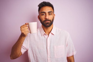 Genç hintli adam gömlek içme kahve fincan ıslanmış pembe arka plan üzerinde akıllı yüz ciddi düşünme emin bir ifade ile ayakta