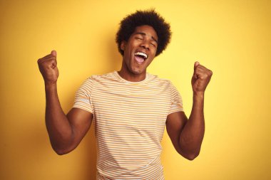 Afro saçlı, çizgili tişört giyen Amerikalı adam izole sarı arka plan üzerinde ayakta kutluyor şaşırmış ve kollarını kaldırdı ve gözleri kapalı başarı için şaşırttı. Kazanan kavramı.