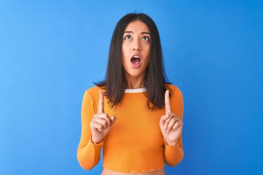 Izole mavi arka plan üzerinde duran turuncu t-shirt giyen genç güzel Çinli kadın şaşırttı ve yukarı bakarak ve parmakları ile işaret ve kolları kaldırdı şaşırttı.
