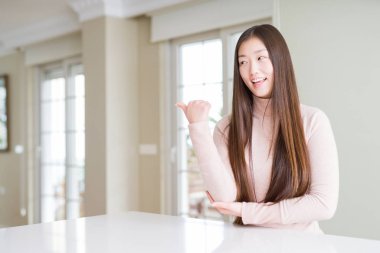 Güzel Asyalı kadın beyaz masada rahat kazak giyen mutlu yüz görünümlü gülümseyerek ve başparmak ile yan işaret.