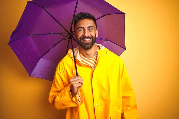 Young indian man wearing raincoat and purple umbrella over isolated yellow background with a happy face standing and smiling with a confident smile showing teeth