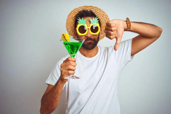 Indian man wearing summer funny look drinking cocktail over isolated white background with angry face, negative sign showing dislike with thumbs down, rejection concept