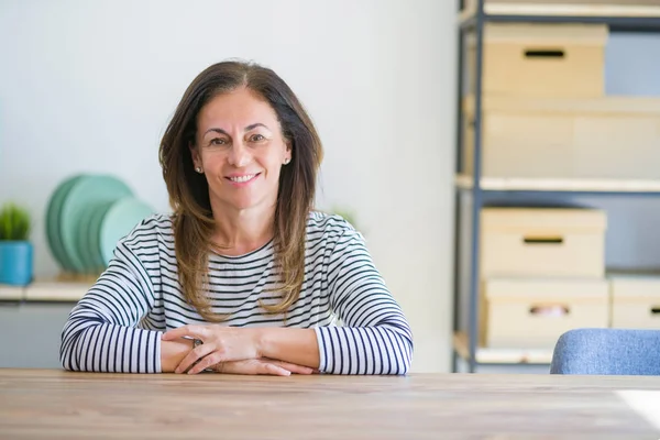Orta yaş kıdemli kadın yüzünde mutlu ve serin bir gülümseme ile evde masada oturuyor. Şanslı kişi.