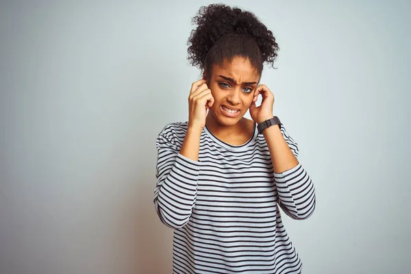 African american woman wearing navy striped t-shirt standing over isolated white background covering ears with fingers with annoyed expression for the noise of loud music. Deaf concept.