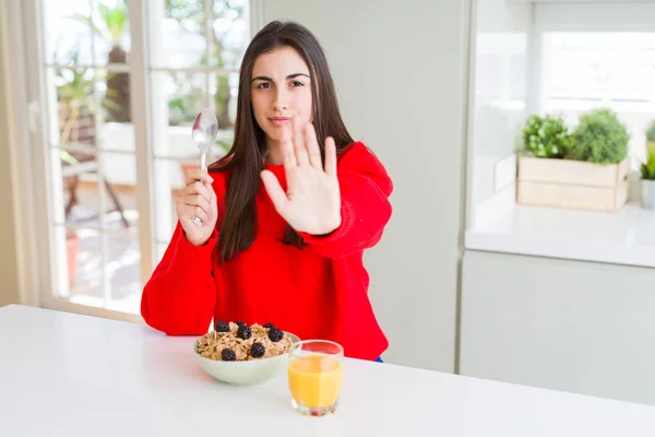Beautiful young woman having healthy cereals and berries for breakfast with open hand doing stop sign with serious and confident expression, defense gesture