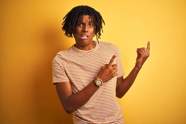 Afro man with dreadlocks wearing striped t-shirt standing over isolated yellow background Pointing aside worried and nervous with both hands, concerned and surprised expression