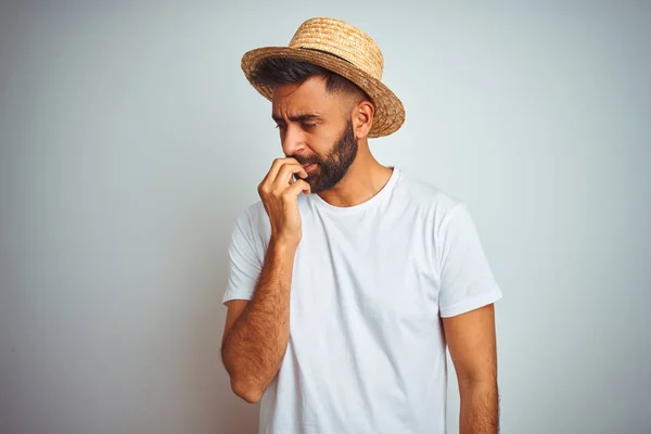 Young indian man on holiday wearing summer hat standing over isolated white background looking stressed and nervous with hands on mouth biting nails. Anxiety problem.