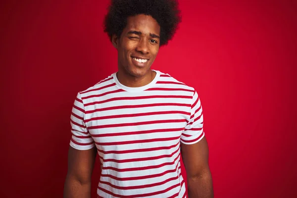 Young african american man with afro hair wearing striped t-shirt over isolated red background winking looking at the camera with sexy expression, cheerful and happy face.