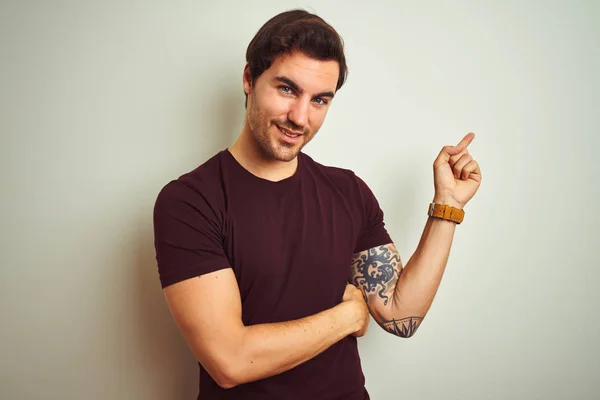 Young handsome man with tattoo wearing purple casual t-shirt over isolated white background with a big smile on face, pointing with hand and finger to the side looking at the camera.
