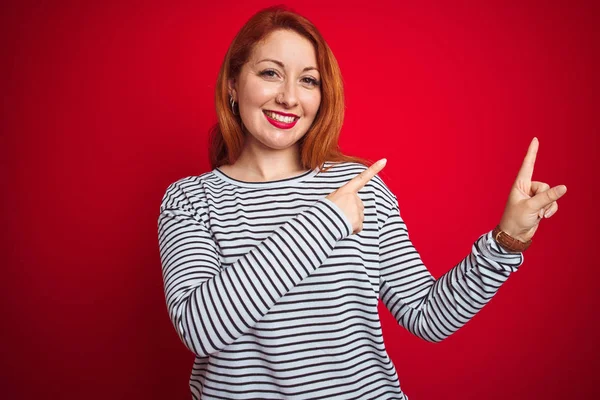 Genç kızıl saçlı kadın kırmızı izole arka plan üzerinde duran kayışlar lacivert gömlek giyen gülümseyerek ve yan iki el ve parmakları ile işaret kamera bakıyor.