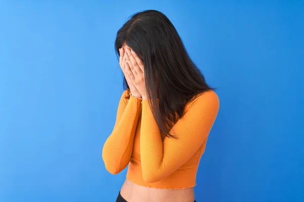 年轻美丽的中国妇女穿着橙色T恤站在孤立的蓝色背景与悲伤的表情覆盖脸与双手 而哭泣 抑郁症概念 — 图库照片