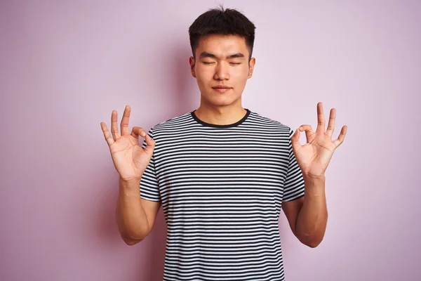 孤立したピンクの背景の上に立つストライプのTシャツを着た若いアジアの中国人男性はリラックスし 指で瞑想ジェスチャーをして目を閉じて微笑んでいます ヨガのコンセプト — ストック写真