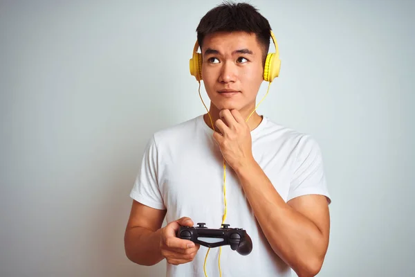 Young asian chinese gamer man playing video game standing over isolated white background serious face thinking about question, very confused idea