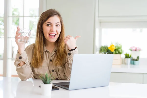 Beautiful young woman working with computer takes a break to drink glass of water pointing and showing with thumb up to the side with happy face smiling