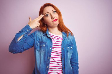 Denim gömlek giyen güzel kızıl saçlı kadın ve izole pembe arka plan üzerinde çizgili t-shirt Çekim ve kendini silah gibi baş el ve parmakları işaret öldürme, intihar jest.