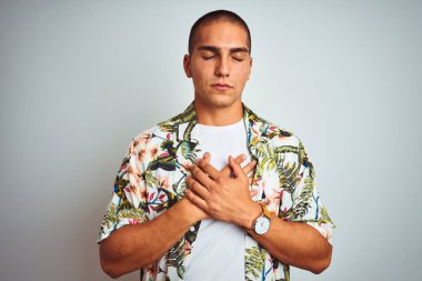 Tatilde beyaz arka plan üzerinde Hawaii gömlek giyen genç yakışıklı adam kapalı gözleri ve yüzünde minnettar jest ile göğüs elleri ile gülümseyerek. Sağlık kavramı.