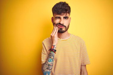 Dövme giyen genç adam diş ağrısı veya diş üzerinde diş hastalığı nedeniyle ağrılı ifade ile el ile el ile ağız dokunmadan izole sarı arka plan üzerinde duran çizgili t-shirt giyiyor. Diş hekimi konsepti.