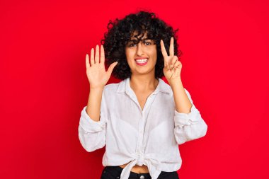 İzole kırmızı arka plan üzerinde beyaz rahat gömlek giyen kıvırcık saçlı genç arap kadın gösteren ve emin ve mutlu gülümserken parmakları ile numara yedi işaret.