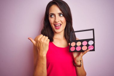 Genç güzel kadın pembe izole arka plan üzerinde makyaj uygulayarak işaret ve mutlu yüz gülümseyerek yan başparmak ile gösteren