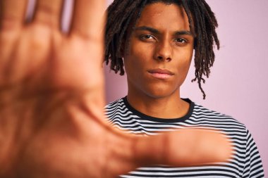 Ciddi ve kendinden emin ifade, savunma jesti ile dur işareti yapıyor açık el ile izole pembe arka plan üzerinde lacivert çizgili t-shirt giyen dreadlocks ile Afro Amerikan adam