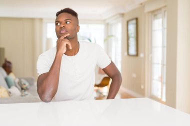 Çene düşünme hakkında soru, dalgın ifade üzerine el ile yakışıklı Afrikalı-Amerikalı adam evde beyaz masada. Düşünceli yüzüyle gülümsüyor. Şüphe kavramı.