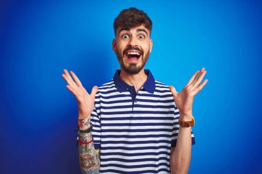 Çizgili polo giyen dövme giyen genç adam izole mavi arka plan üzerinde duran deli kutluyor ve kollarını kaldırdı ve açık gözleri heyecan çığlık ile başarı için hayran. Kazanan kavramı