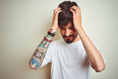 Dövmeli genç adam izole beyaz arka plan üzerinde ayakta t-shirt giyen baş ağrısı umutsuz muzdarip ve stresli çünkü ağrı ve migren. Eller baş üstünde.