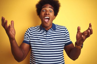 Afro saçlı amerikan adam lacivert çizgili polo giyen izole sarı arka plan üzerinde duran silah kaldırdı ve kapalı gözleri heyecan çığlık ile başarı için deli ve deli kutluyor. Kazanan kavramı